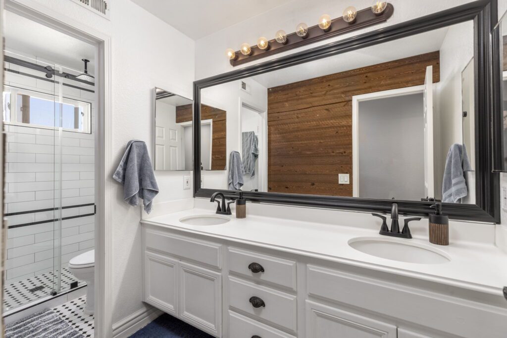 Modern bathroom featuring a double vanity with black fixtures, framed mirrors, and a walk-in shower with white tile. Warm wood accent wall adds contrast.