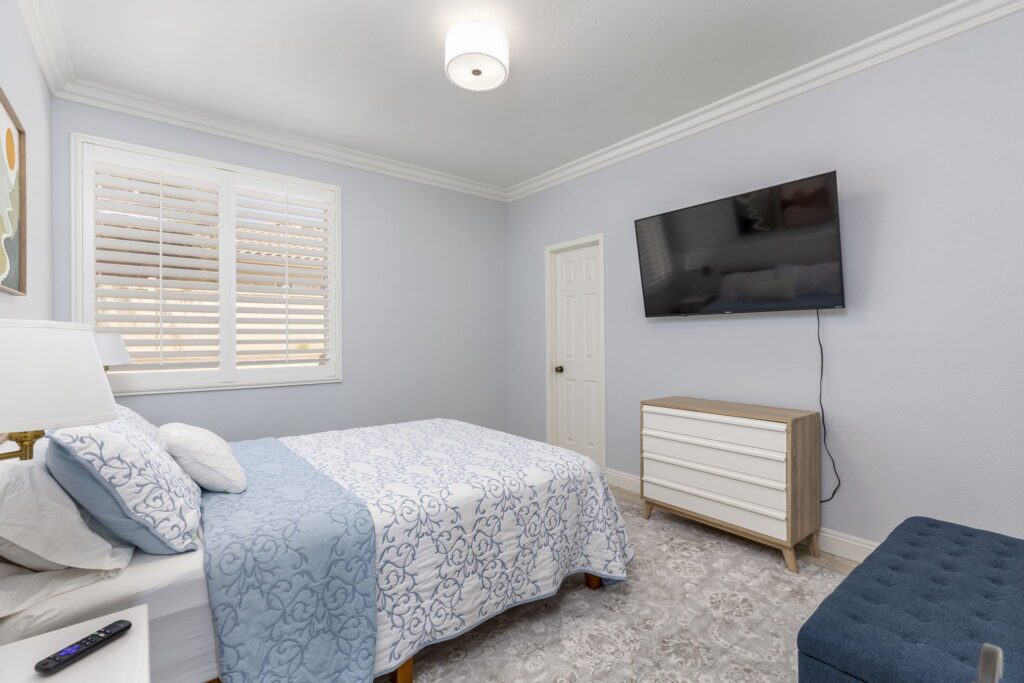 Cozy bedroom featuring a queen bed with blue and white bedding, a mounted TV, and a soft rug. Natural light filters through window shutters.