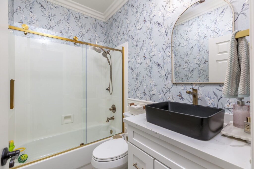 Bright, modern bathroom featuring floral wallpaper, a sleek black sink, and a glass shower with a gold-framed sliding door.