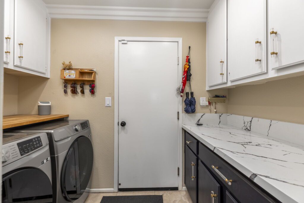 Modern laundry room featuring gray washing machines, white cabinets, and a marble countertop, with a door leading to another space.