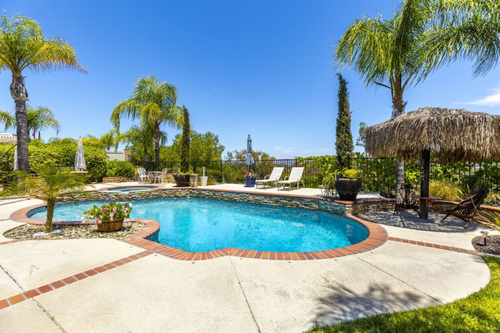 Lush backyard pool area featuring a sparkling blue pool surrounded by palm trees, lounge chairs, and a thatched umbrella for relaxation.