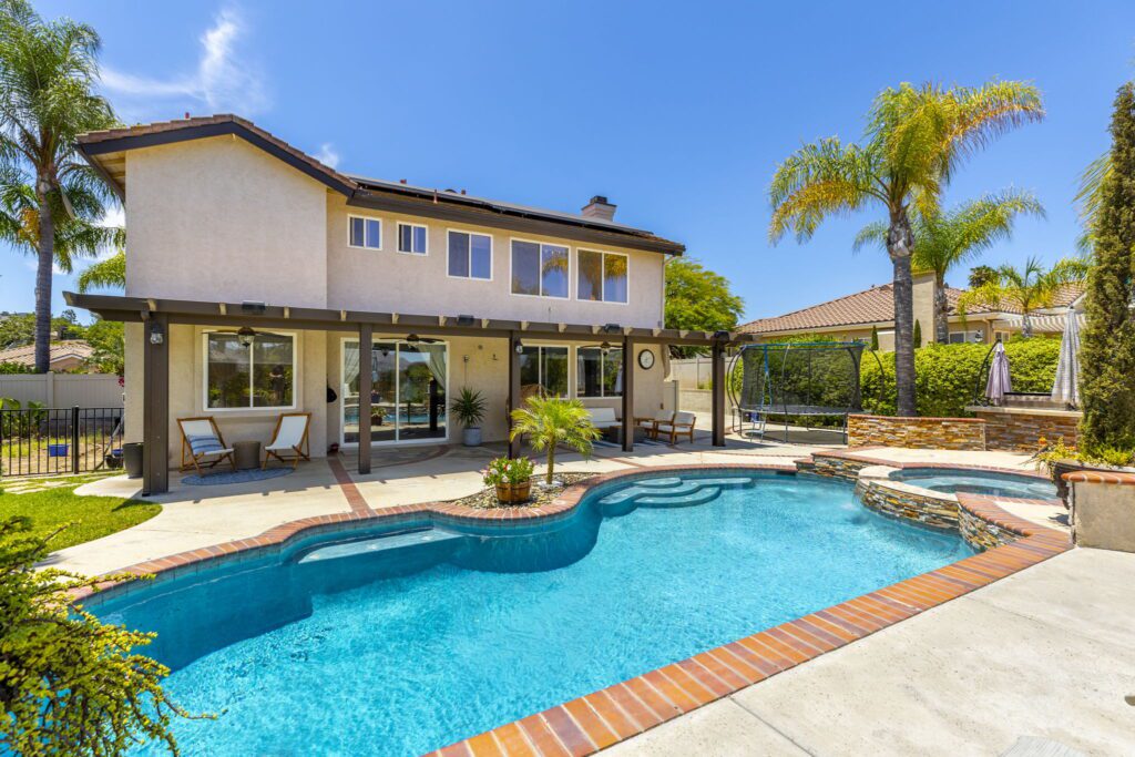 Modern two-story house with a swimming pool and patio area. Lush palm trees and a swing set enhance the backyard’s inviting atmosphere.