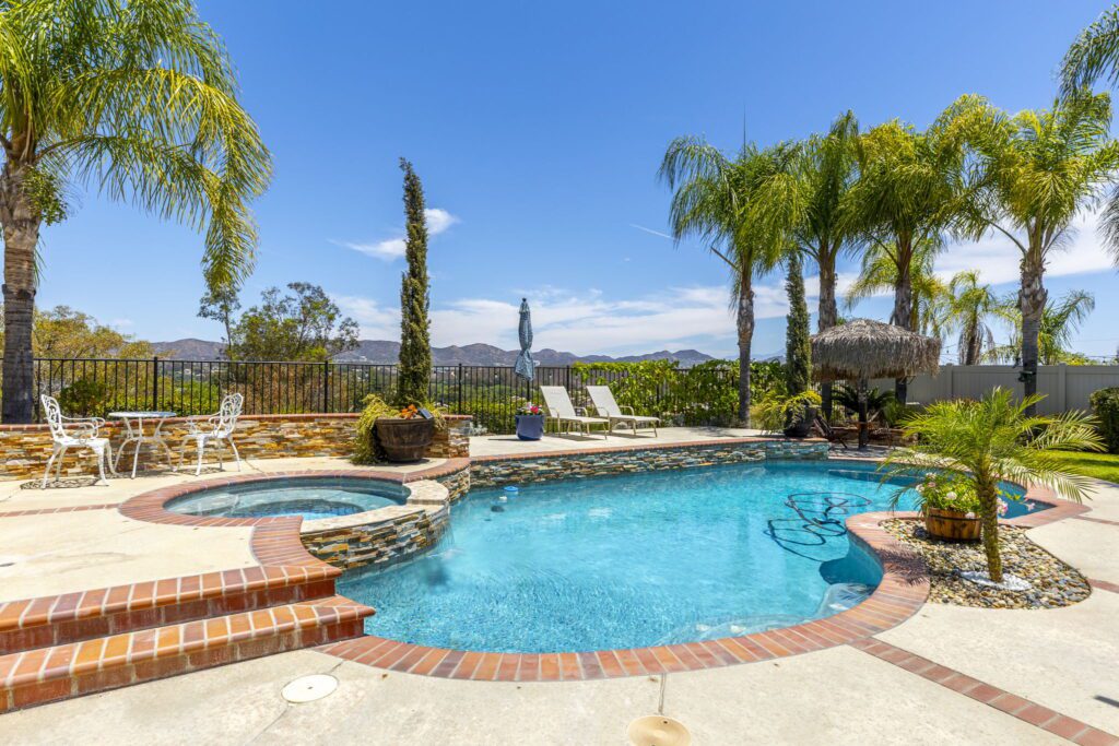 Inviting pool area featuring a sparkling blue pool surrounded by palm trees, lounge chairs, and a shaded seating area, set against a clear blue sky.