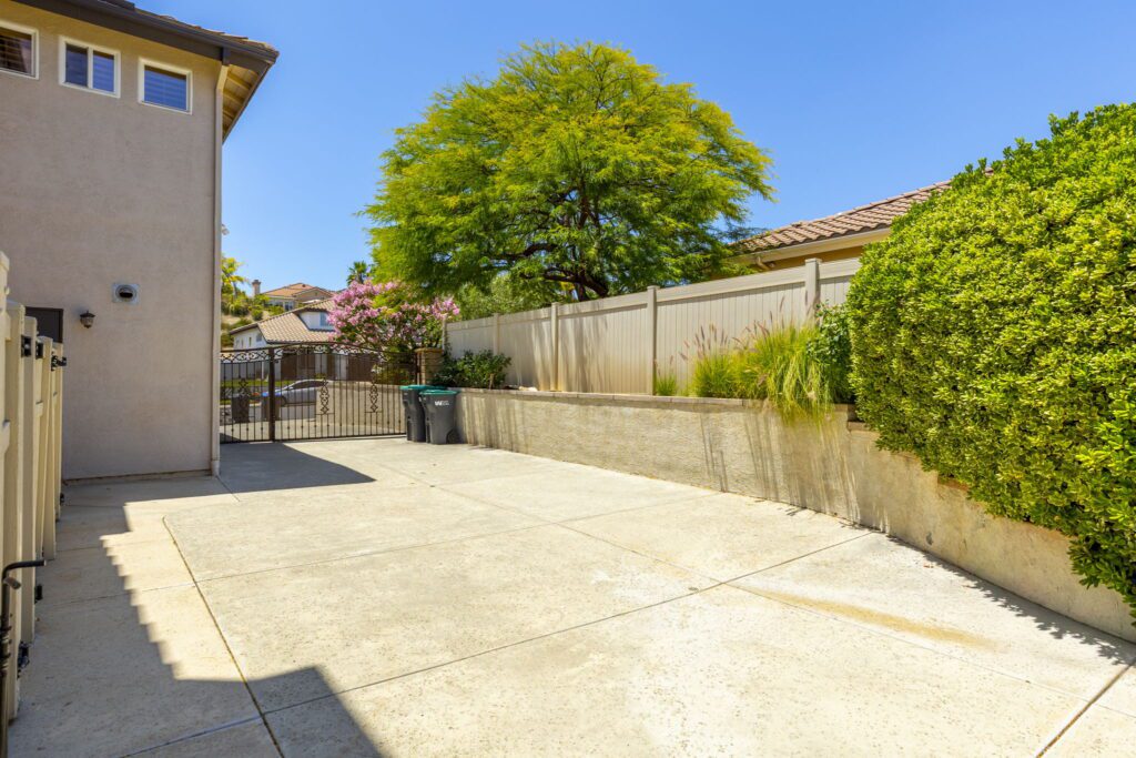 Spacious outdoor area with a concrete driveway, a green trash bin, and lush plants, bordered by a fence and a house wall, under clear blue skies.