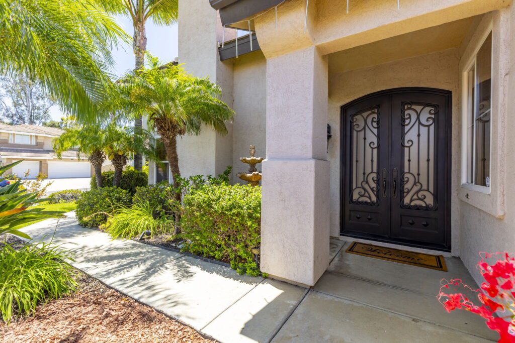 Elegant entryway featuring ornate black double doors and a welcoming mat. Lush tropical plants and a small fountain enhance the inviting atmosphere.
