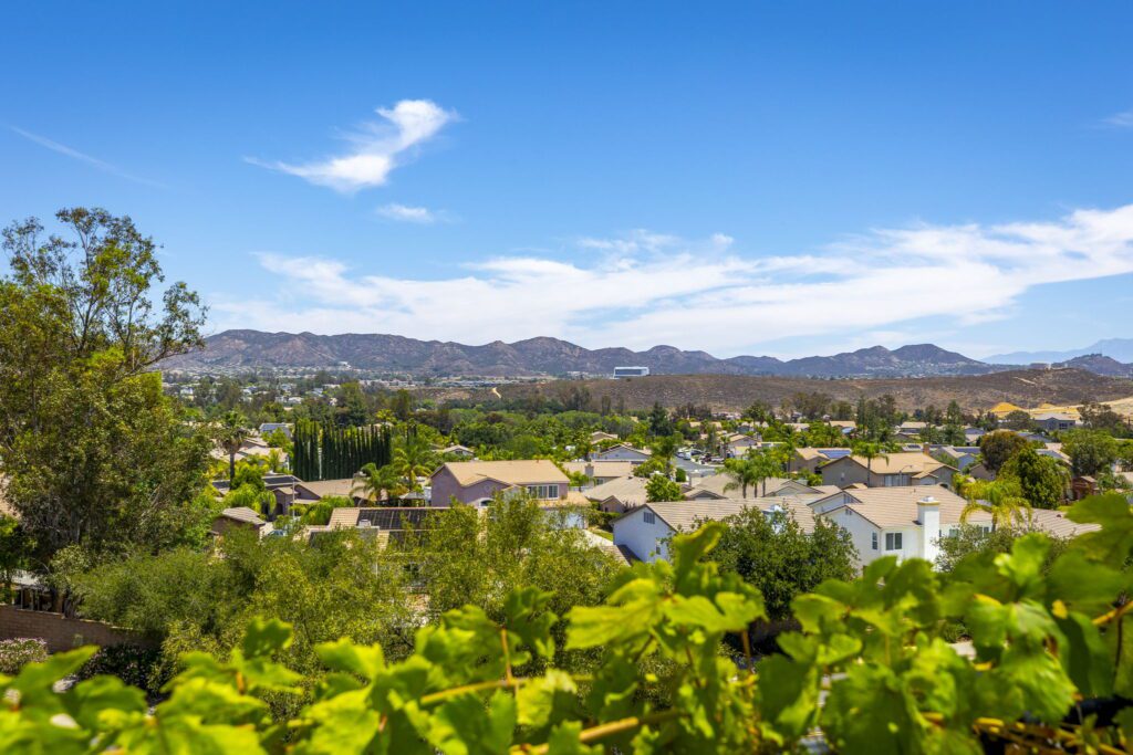 Expansive view of suburban rooftops with greenery in the foreground and rolling mountains under a clear blue sky, showcasing a peaceful landscape.