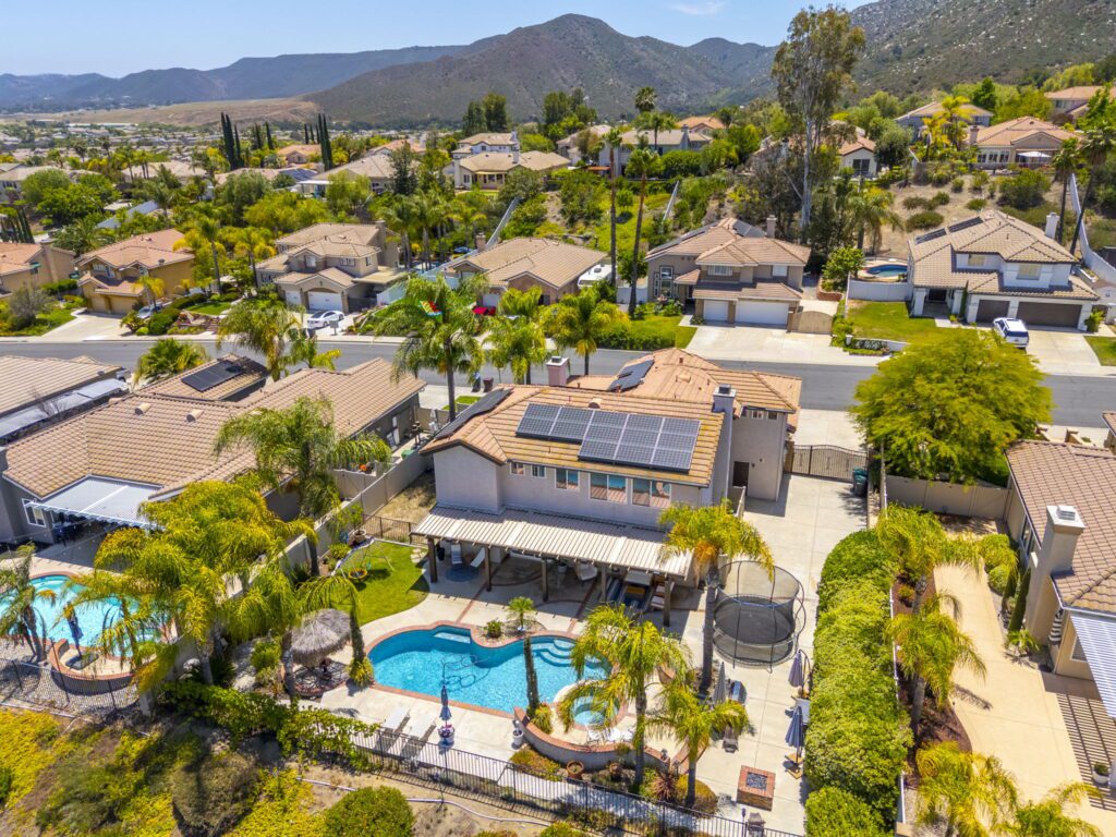 Aerial view of a suburban home with solar panels, featuring a backyard pool, palm trees, and nearby houses against a mountainous backdrop.