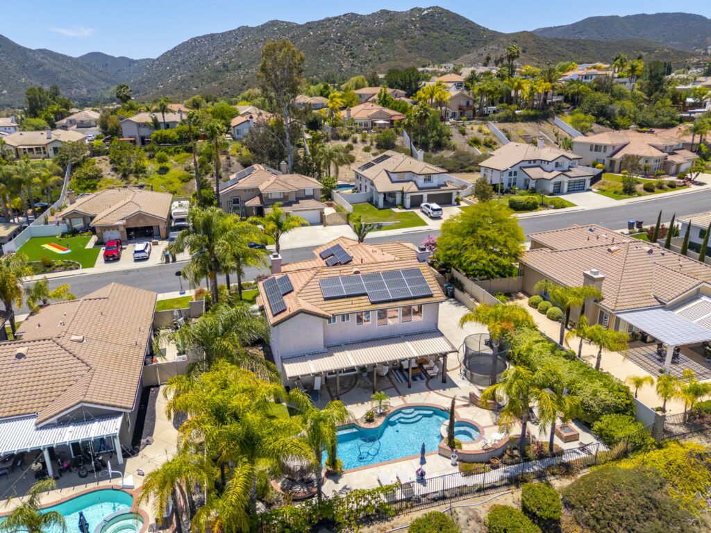 Aerial view of a suburban neighborhood featuring houses with solar panels, a pool, and palm trees, set against a backdrop of rolling hills.