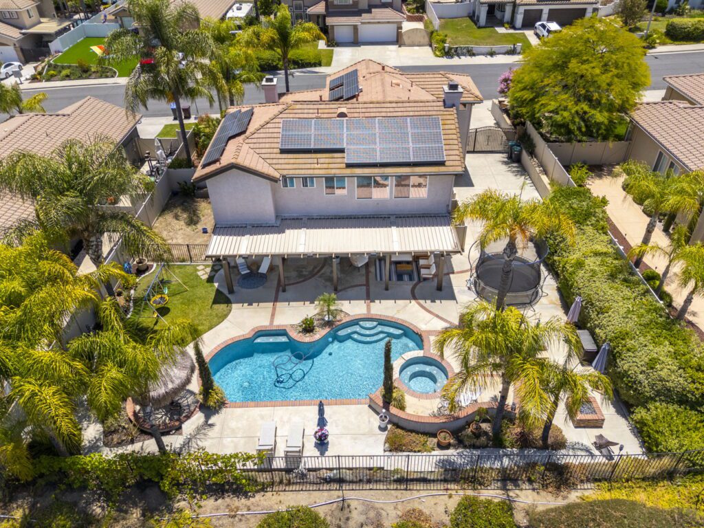 Aerial view of a residential backyard featuring a swimming pool, spa, patio area with seating, and solar panels on the roof of the house.
