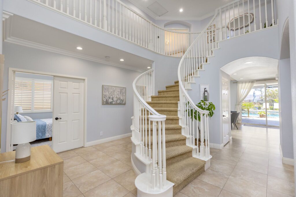 Bright, elegant staircase with white railings leads to a second floor, framed by light walls and large windows showcasing a pool view.