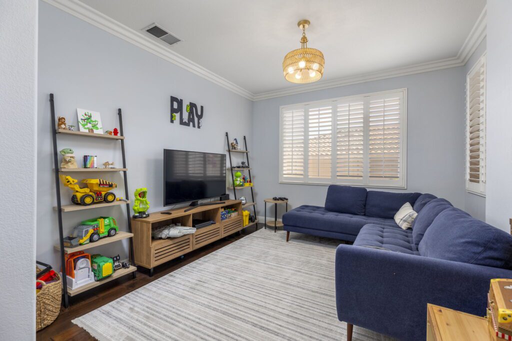 Bright playroom featuring a blue sectional sofa, toys on shelves, and a TV stand. Decor includes a playful "PLAY" sign and colorful decor.