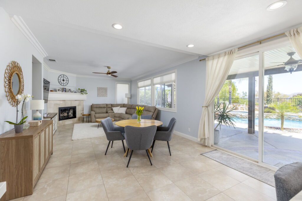 Bright living area featuring a circular dining table and a cozy sectional sofa. Large windows overlook a pool area, enhancing a casual, inviting atmosphere.