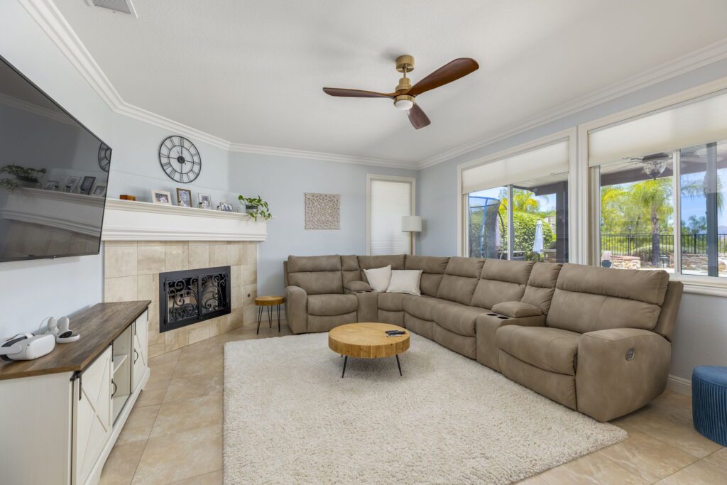 Cozy living room featuring a large sectional sofa, a wooden coffee table, and a TV mounted above a fireplace. Sunlight streams through large windows.
